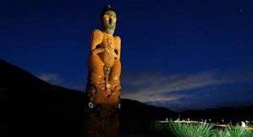Statue of Umukaria, a Māori carving illuminated at night against a dark sky, with surrounding greenery subtly visible in the foreground