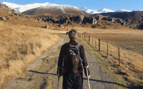 A man walks down a gravel road towards some hills, he is using crutches 