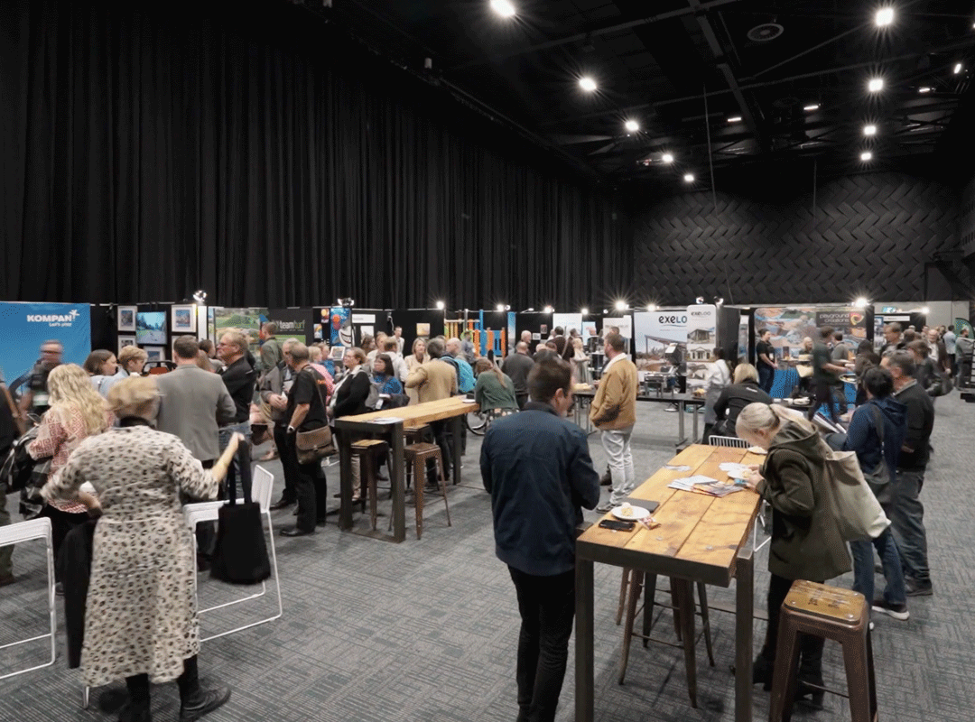 People interacting at a trade booth area, viewing displays and engaging with representatives. The space is lively, with various booths showcasing products and services