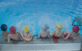 A group of children wearing brightly coloured swim caps and swimsuits sit on the edge of a swimming pool, their feet splashing in the water.