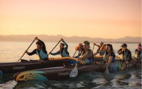 A group of people sit in a double-hulled waka (canoe) in the water. They are holding paddles and rowing. Three people sit on each side.