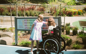 Two people look at a communication board in a playground. A young girl points at the board while a lady in a wheelchair reads it